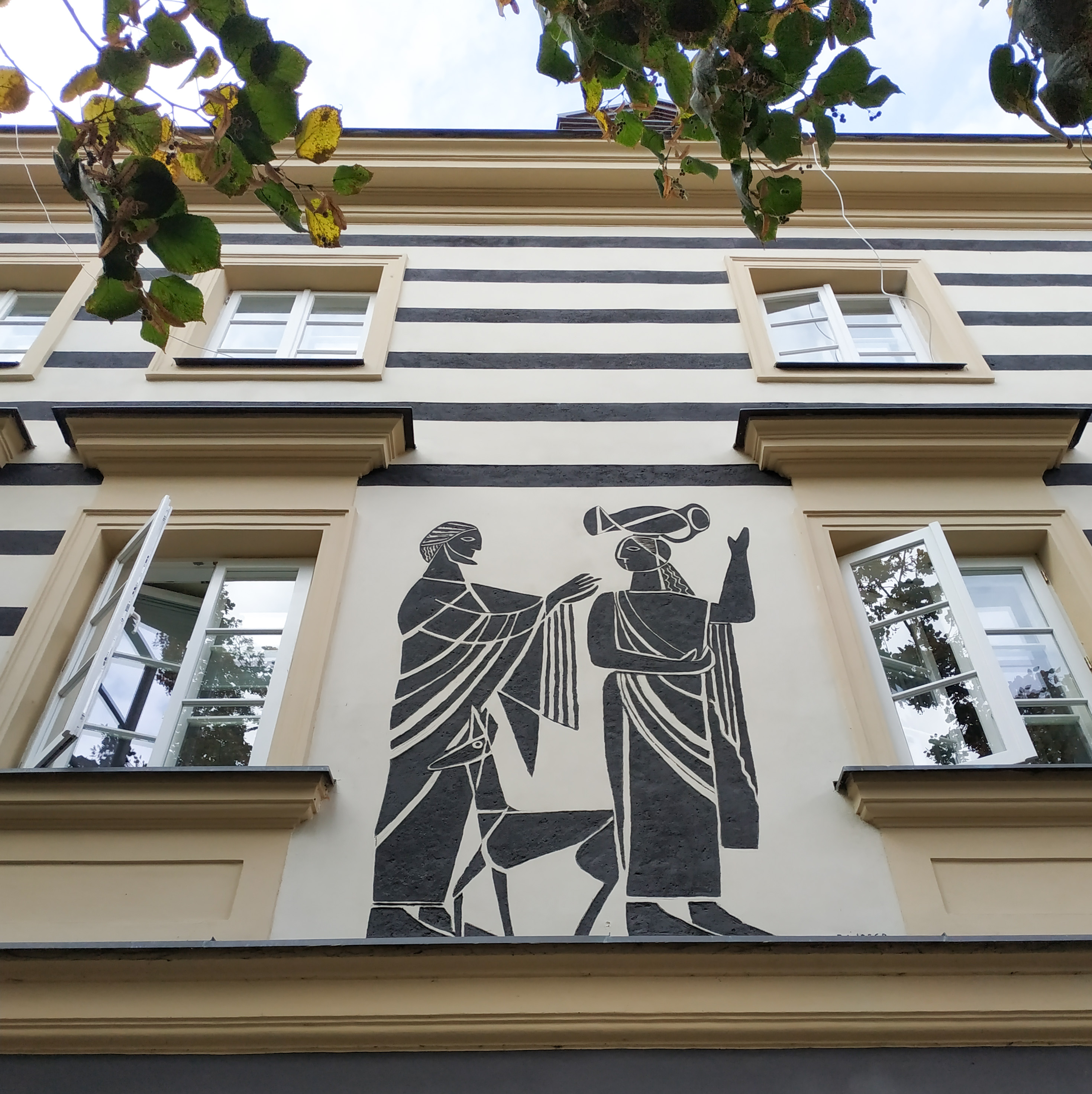 ściana budynku w kolorze kremowymz oknami, na jej tle w kolorze czarnym  fresa przestawiajaca kobietę z dzbanem na głowie i mężczyznę z psem 